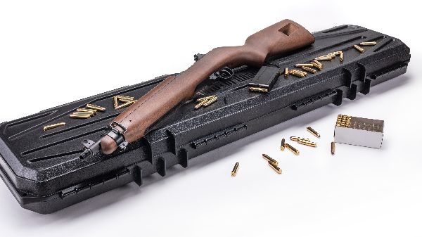 A rifle with a wooden stock rests on a black hard case, surrounded by scattered ammunition and an open box of bullets on a white surface.