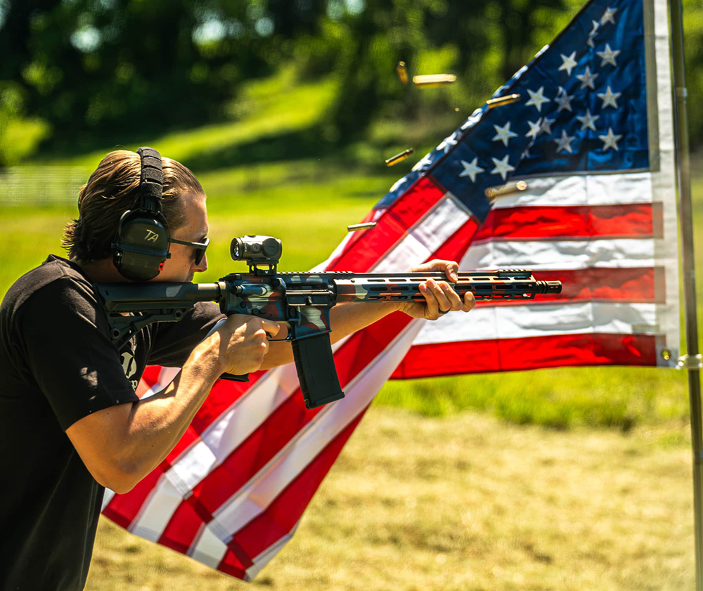 Person wearing ear protection fires a rifle outdoors with spent bullet casings visible in the air; an American flag is displayed in the background.