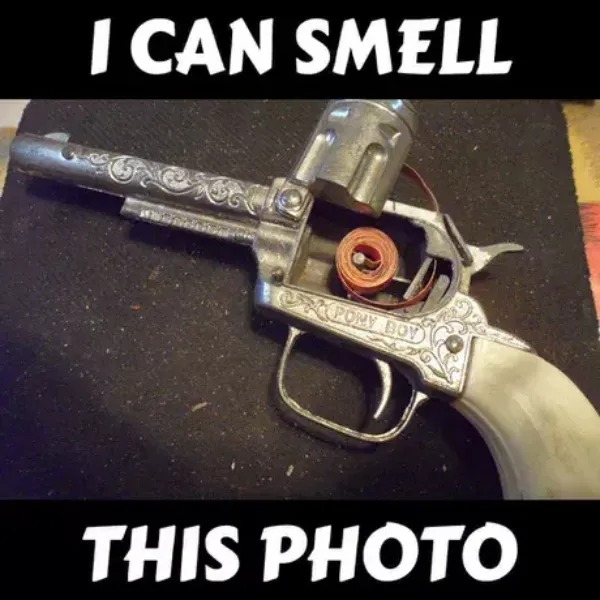 A vintage cap gun with decorative engravings and a roll of caps inside, placed on a dark surface. Text above and below reads, "I CAN SMELL THIS PHOTO.