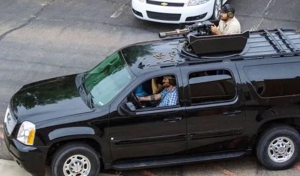 A black SUV with an open roof hatch shows one person aiming a mounted firearm while others sit inside. Live ammunition casings are scattered on the roof.