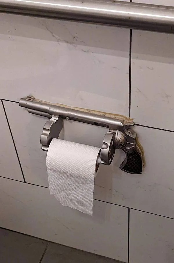 A toilet paper holder made from a revolver is mounted on a tiled bathroom wall, holding a roll of toilet paper.