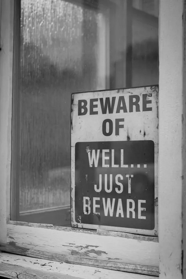 A weathered sign in a window reads, “BEWARE OF WELL... JUST BEWARE.” The image is in black and white.