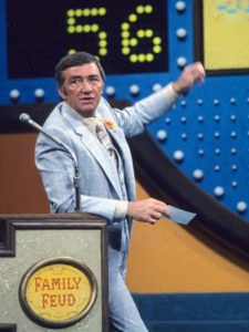 A man in a light gray suit gestures toward a scoreboard while standing at a podium labeled “Family Feud.”.